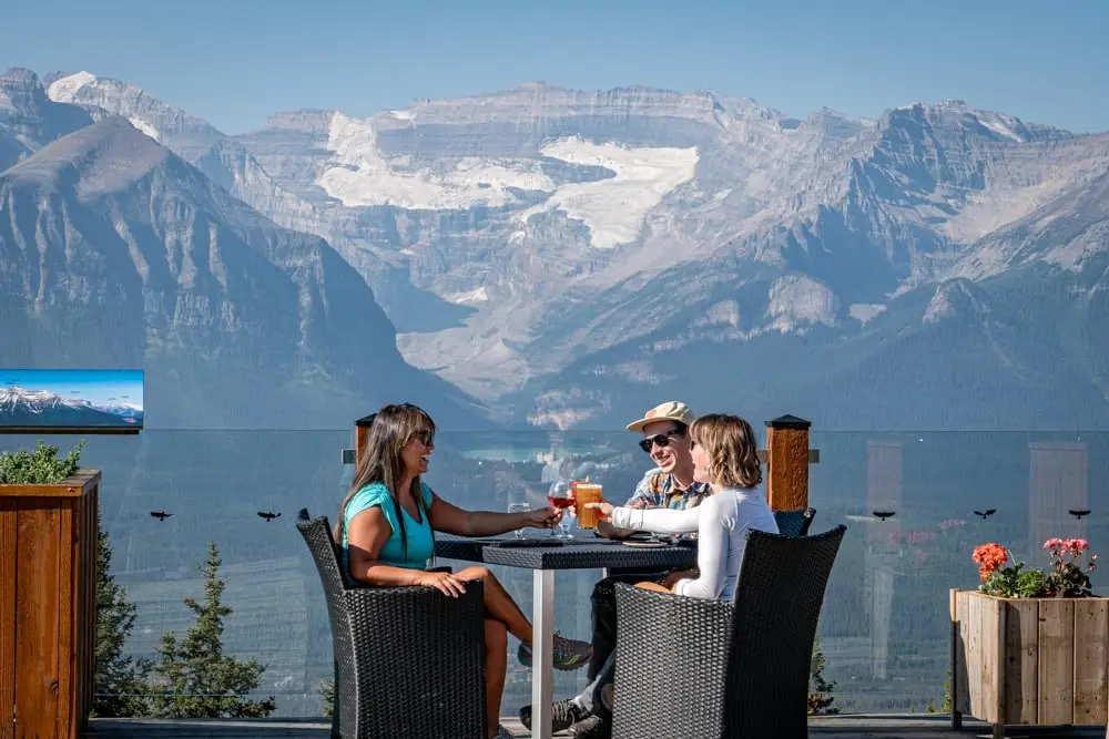 Friends toast drinks on an alpine bistro patio with lake and glaciers in the background.