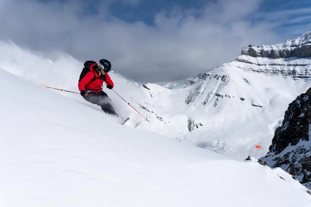 Skier in a red jacket carves a steep powder slope with spindrift.