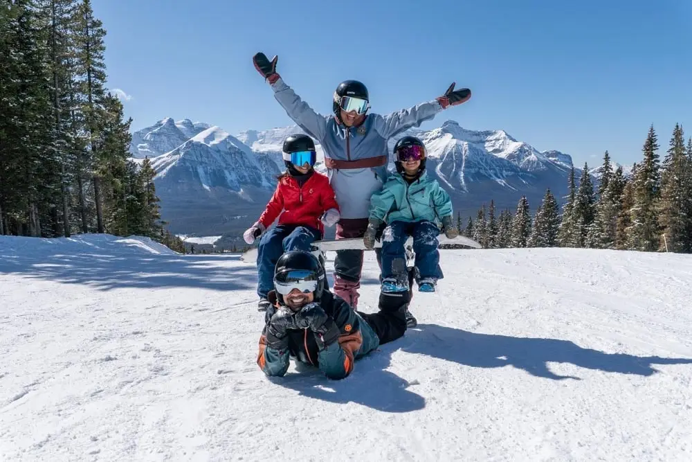 Family poses on a groomed ski run with mountain peaks in the background.