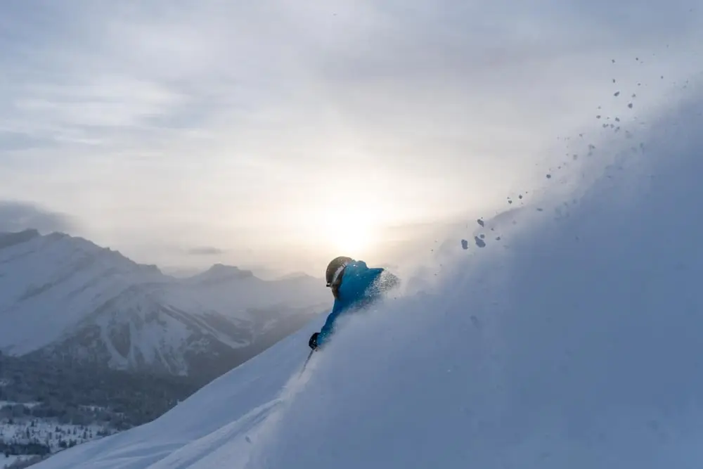 Skier carves a sunrise powder turn with snow backlit against alpine peaks.