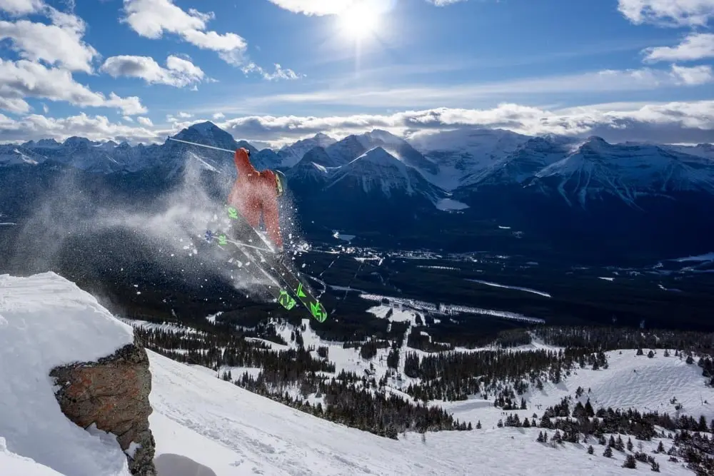 Skier launches a cliff drop with spray, valley, and mountain peaks behind.