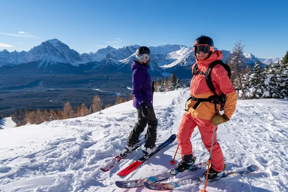 Two skiers pause on a sunny ridge with panoramic mountain peaks behind them.