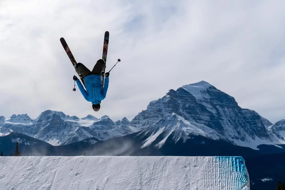 Freestyle skier performs an inverted flip off a jump with mountains behind.